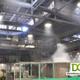 Oświetlenie przemysłowe hali fabrycznej z widocznymi lampami i konstrukcją stalową pod sufitem. W tle unosząca się para. DGE oświetlenie dla przemysłu.
