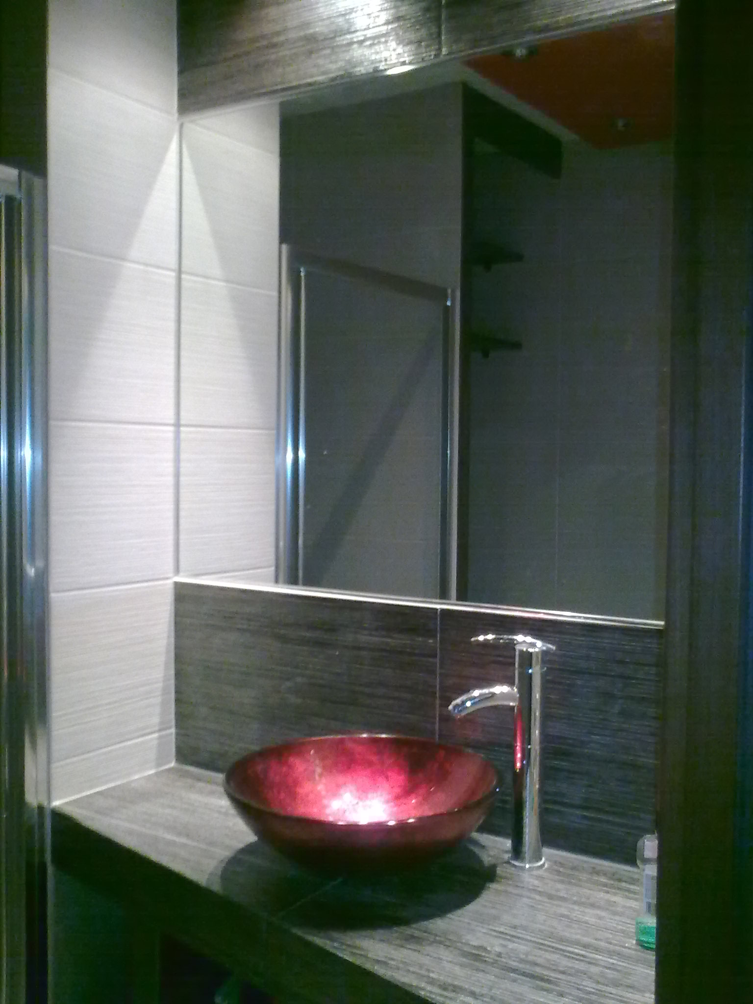 Umywalka z czerwonego szkła i wysoka chromowana bateria w nowoczesnej łazience z szarymi płytkami imitującymi drewno i lustrem. Widoczny fragment kabiny prysznicowej.