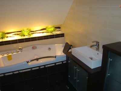 Nowoczesna łazienka na poddaszu z wanną jacuzzi i umywalką nablatową na szafce w ciemnej kolorystyce, oświetlona listwą z roślinami dekoracyjnymi.