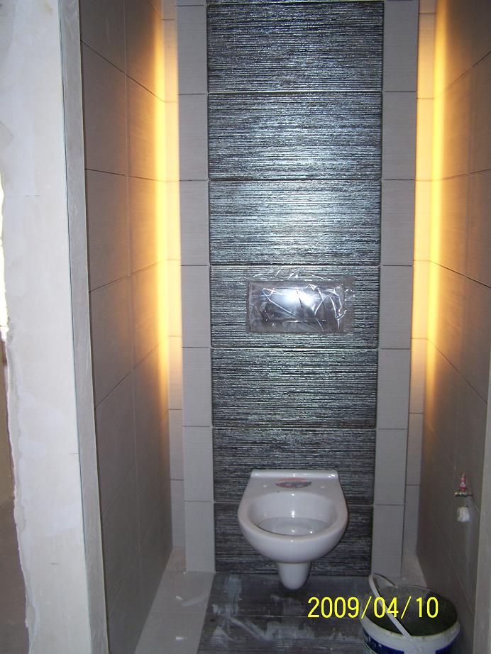 Wnętrze małej łazienki z podwieszaną toaletą na tle dekoracyjnej ściany z płytek o fakturze drewna, oświetlonej bocznym światłem. Widoczne ślady prac wykończeniowych i wiadro na podłodze.