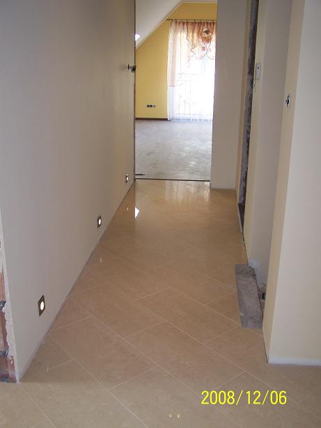 Wykończony korytarz z beżową, ułożoną w jodełkę podłogą z płytek, oświetleniem punktowym w ścianie i widokiem na pokój z żółtymi ścianami i jasnymi zasłonami.