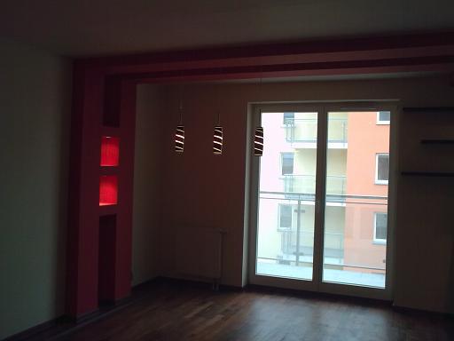 Pusty pokój z parkietem, czerwonymi elementami dekoracyjnymi na ścianach, trzema wiszącymi lampami i dużym oknem balkonowym z widokiem na budynek mieszkalny.