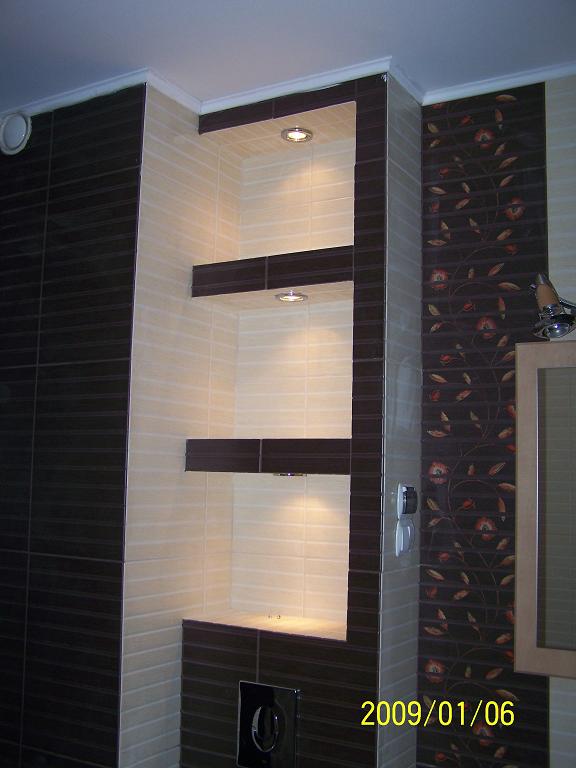 Półki wnękowe z podświetleniem LED, wykończone brązowymi i beżowymi płytkami w łazience z dekoracyjną ścianą w kwiatowy wzór. Widoczny panel sterowania toalety podwieszanej.