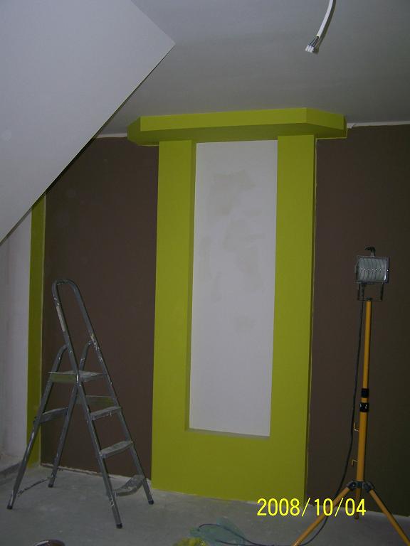 Wnętrze w trakcie prac wykończeniowych: fragment ściany pomalowany na brązowo z elementem dekoracyjnym w kolorze limonkowym, drabina malarska i lampa halogenowa na statywie.