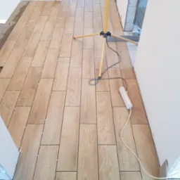 Układanie podłogi z płytek imitujących drewno w korytarzu, widoczne krzyżyki dystansowe, lampa budowlana i przedłużacz.