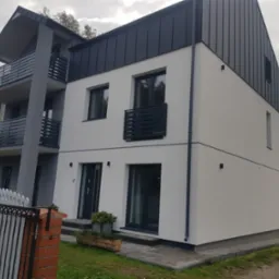 Nowoczesny dwupiętrowy dom z szarą blachą na dachu i białą elewacją, z balkonami z szarymi balustradami, widziany z perspektywy ogrodu.