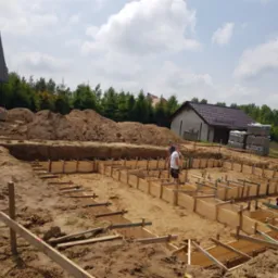 Przygotowanie szalunków pod fundamenty domu jednorodzinnego na piaszczystym podłożu, widoczne zbrojenia i wykop, w tle budynek w stanie surowym otwartym oraz sterta ziemi.