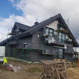 Budowa domu jednorodzinnego: czarny styropian na elewacji, rusztowania, pracownik na wysokości, częściowo otynkowana ściana, biały bus w tle.