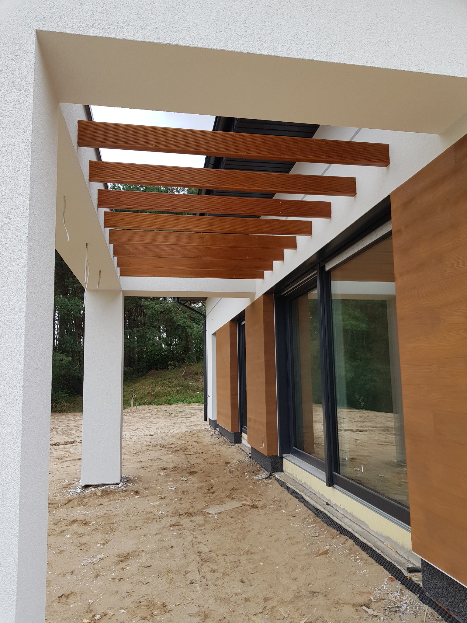 Nowoczesny dom z drewnianą pergolą nad tarasem i elewacją wykończoną panelami imitującymi drewno, widok z zewnątrz na surowym terenie.