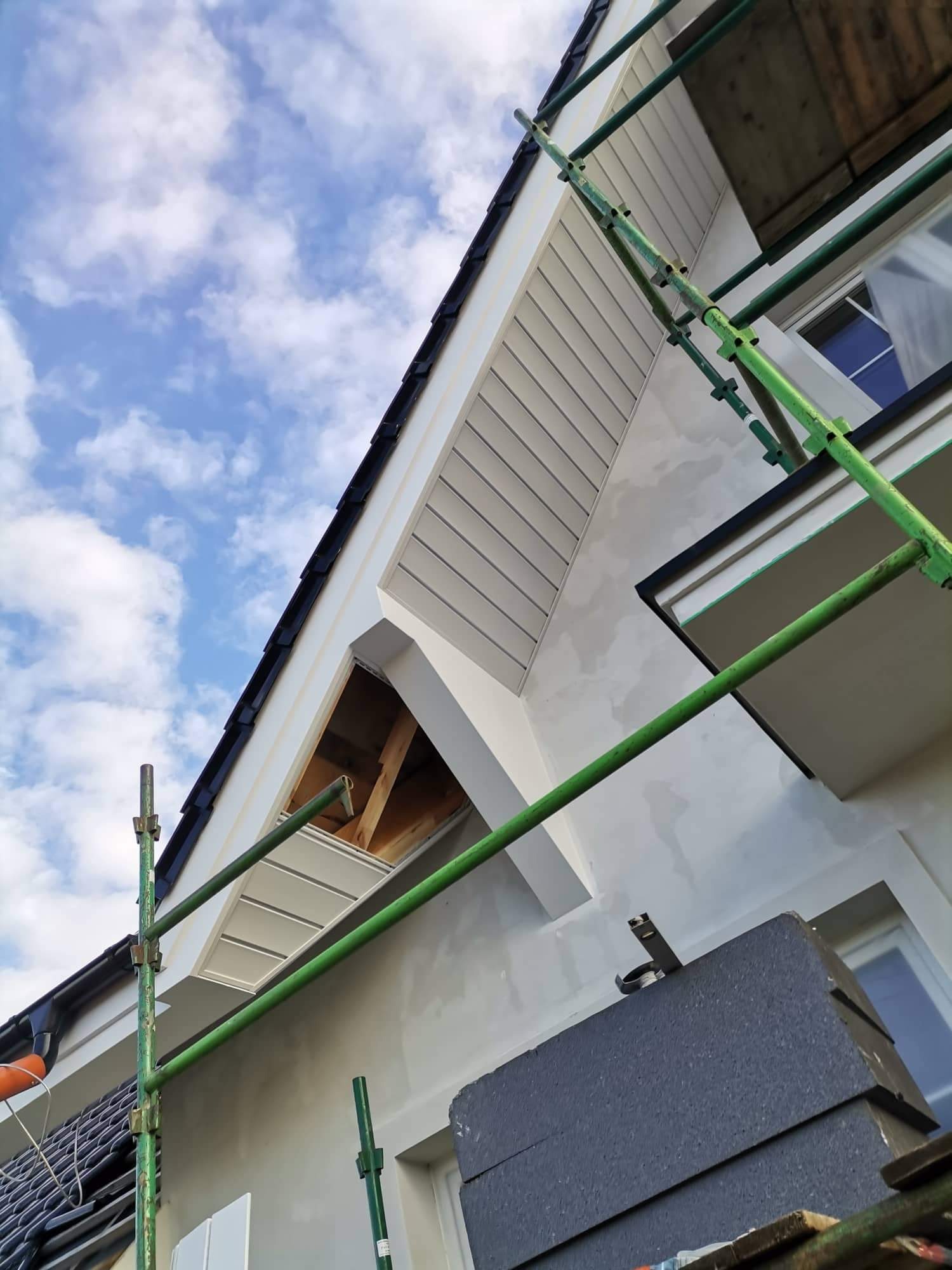 Budynek w trakcie prac elewacyjnych, widoczne rusztowanie, niezakończona powierzchnia ściany, fragment dachu z czarną dachówką oraz odsłonięta konstrukcja poddasza.