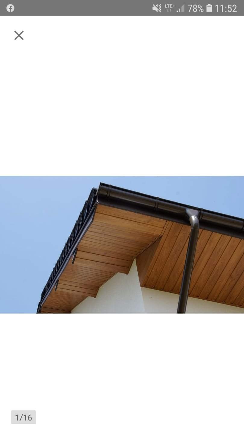 Drewniana podbitka dachowa z czarną rynną i rurą spustową na tle błękitnego nieba, widok z dołu.