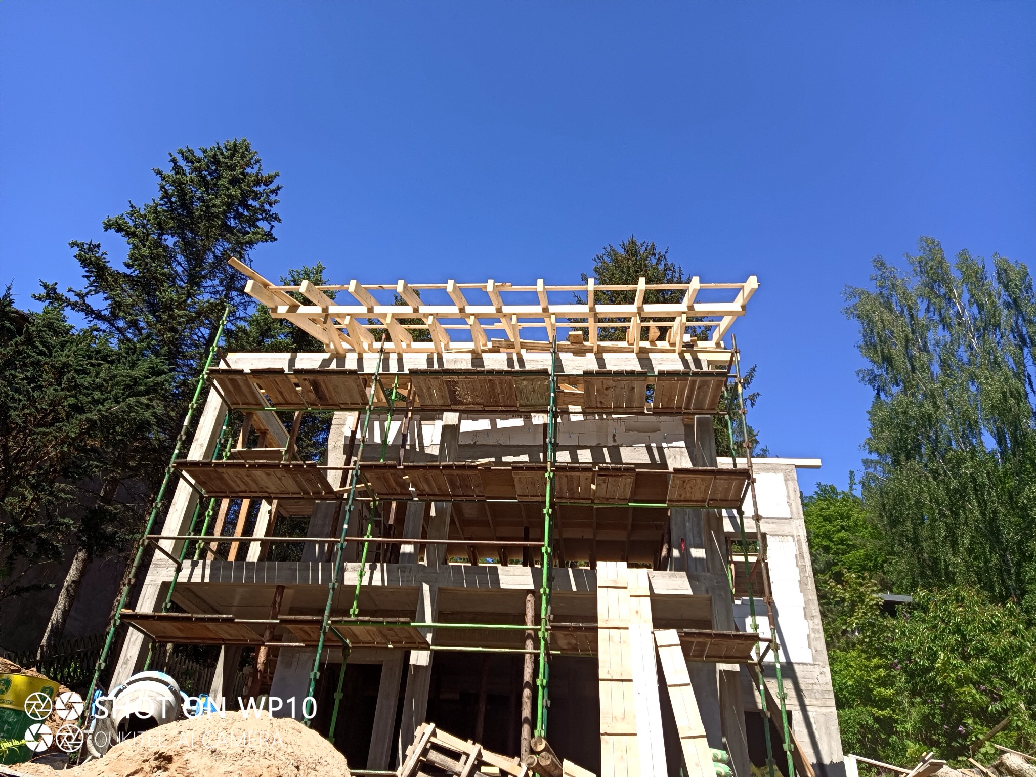 Wznoszenie szkieletu dachu na surowym, dwukondygnacyjnym domu z betonu komórkowego, widoczne rusztowania i błękitne niebo.