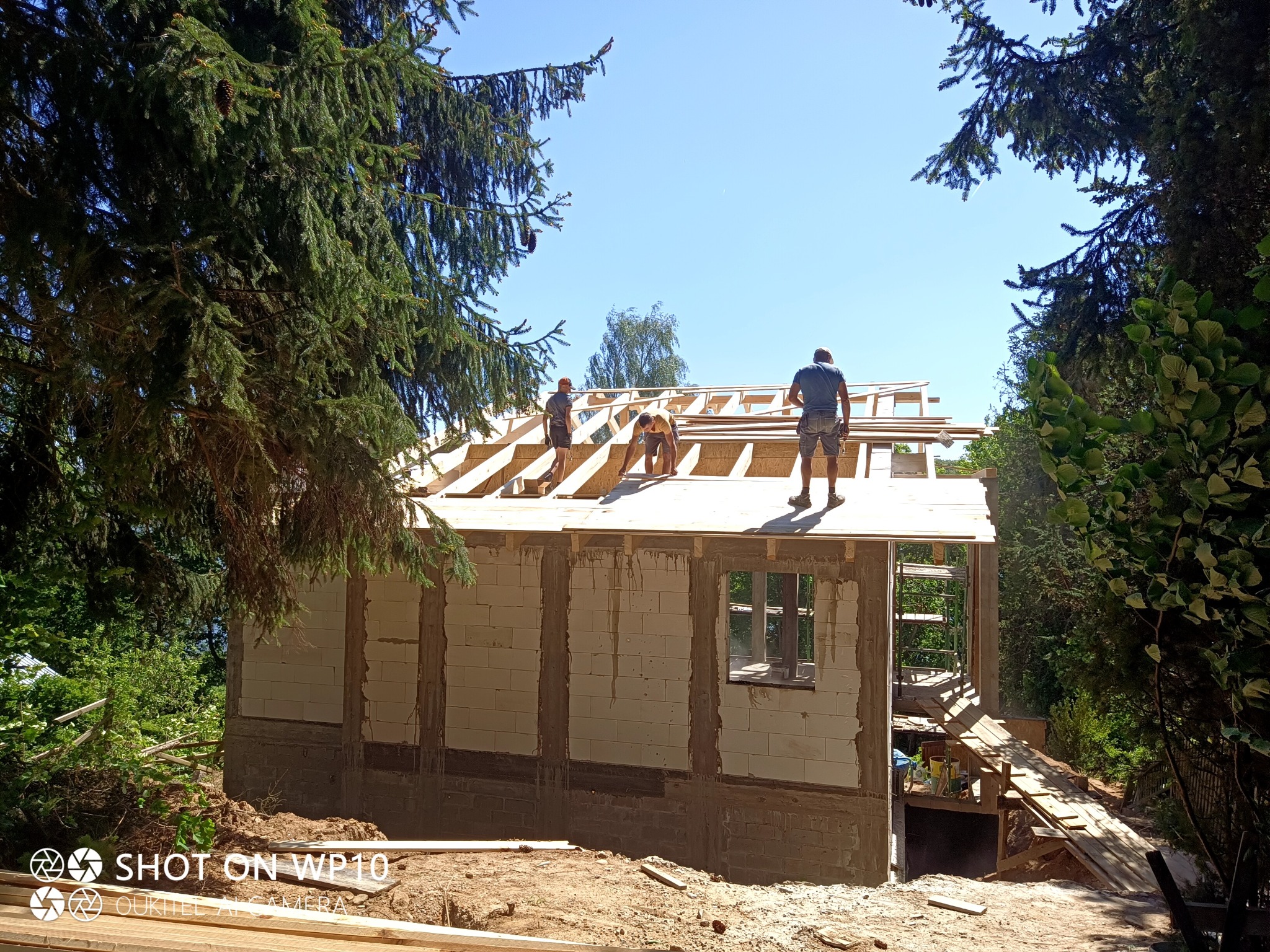 Budowa domu jednorodzinnego z białych bloczków, trzech robotników pracuje na drewnianej konstrukcji dachu, widok z poziomu gruntu w otoczeniu zieleni.