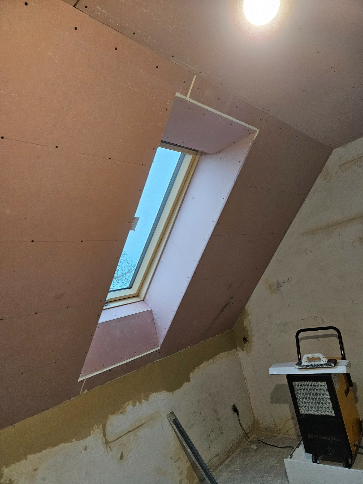 Płyty gipsowo-kartonowe na poddaszu z oknem dachowym, widoczne ślady po szpachlowaniu. Grzejnik elektryczny w rogu pomieszczenia. Prace wykończeniowe.