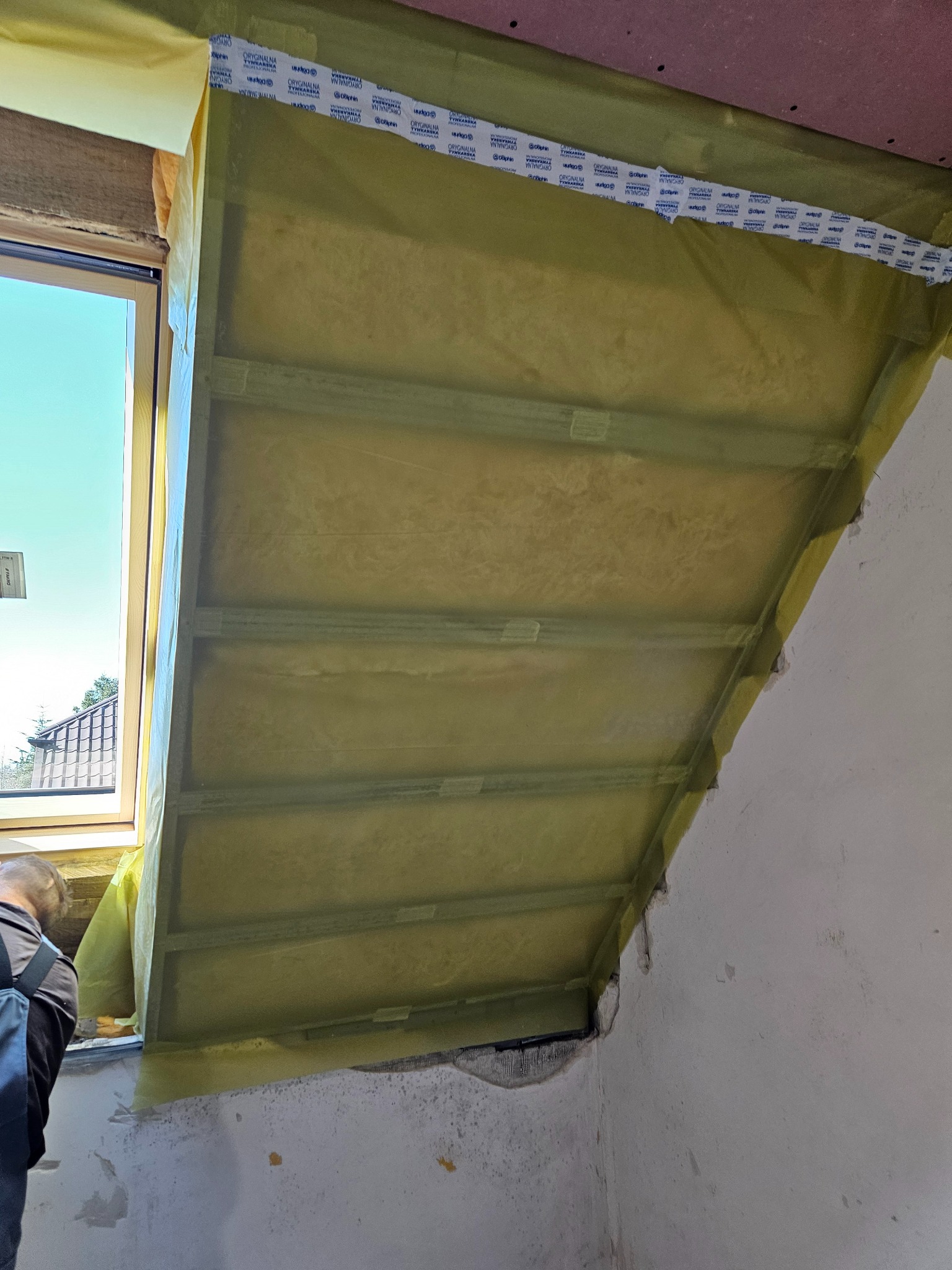 Ocieplenie poddasza wełną mineralną, widoczne profile i folia paroizolacyjna przy oknie dachowym. Prace wykończeniowe w trakcie realizacji.