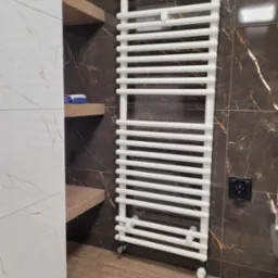 Biały grzejnik łazienkowy na tle ciemnobrązowych płytek imitujących marmur, z widocznym gniazdkiem elektrycznym i drewnianymi półkami z kosmetykami.