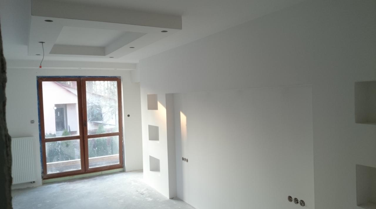 Jasne wnętrze po remoncie: białe ściany z dekoracyjnymi wnękami, drewniane okno i sufit podwieszany z oświetleniem, widok na zewnątrz.