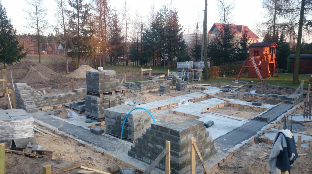Fundamenty domu w trakcie budowy, częściowo wymurowane z bloczków betonowych, widoczne szalunki i wykop, w tle drzewa i dom sąsiada.