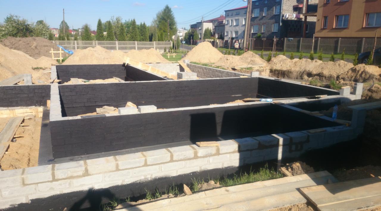 Fundamenty domu w trakcie budowy: izolowane ściany z bloczków, przygotowane do dalszych prac, z widocznymi hałdami ziemi w tle.