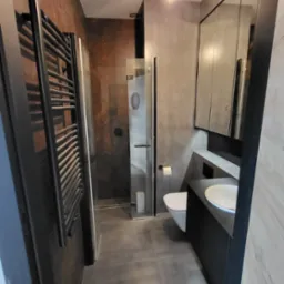 Nowoczesna łazienka z kabiną prysznicową, grzejnikiem drabinkowym w kolorze czarnym, umywalką z szafką i lustrem w zabudowie oraz toaletą, utrzymana w szaro-brązowej kolorystyce.