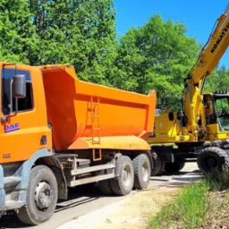 Pomarańczowa wywrotka DAF CF i żółta koparka Komatsu pracują obok wykopu z widoczną niebieską rurą, w tle zieleń drzew i zaparkowane samochody.