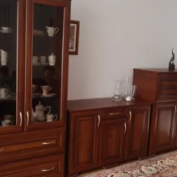 Komplet mebli kuchennych w kolorze ciemnego drewna z przeszkloną witryną wypełnioną porcelaną i dekoracjami, ustawiony na tle jasnej ściany i wzorzystego dywanu.