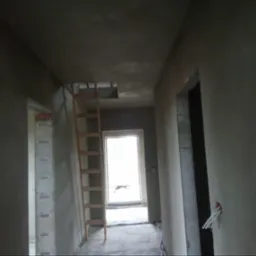 Korytarz w trakcie prac wykończeniowych: ściany po nałożeniu tynku maszynowego, widoczna drewniana drabina oparta o ścianę, przygotowane otwory drzwiowe, wystające przewody elektryczne.