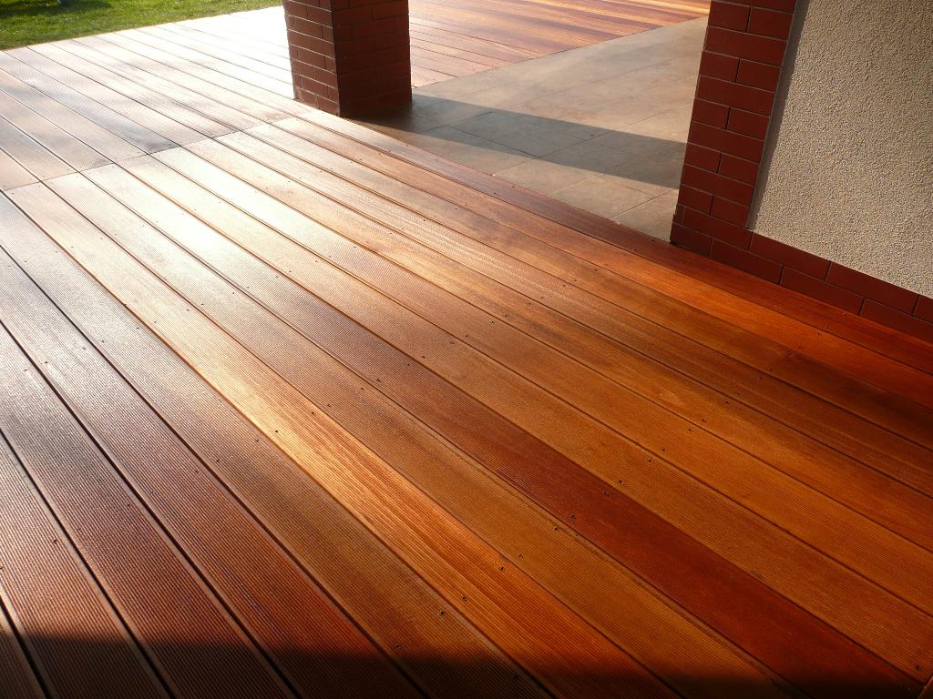 Egzotyczny taras drewniany z desek ułożonych równolegle, z widocznymi wkrętami, przylegający do muru z cegły i fragmentu ściany o szarej strukturze.