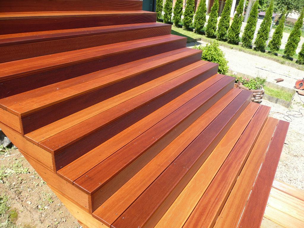 Schody zewnętrzne wykonane z egzotycznego drewna o ciepłych odcieniach brązu, prowadzące do ogrodu z tujami.