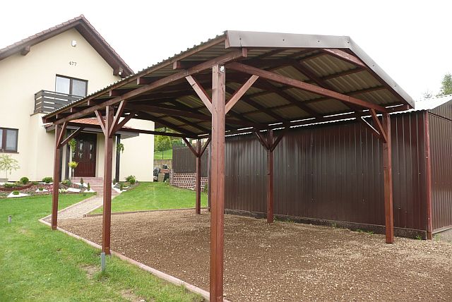 Drewniana wiata garażowa z metalowym dachem, wsparta na solidnych słupach, na tle domu jednorodzinnego i brązowego ogrodzenia.