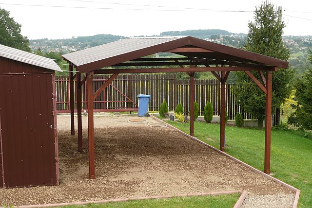 Drewniana wiata garażowa z metalowym dachem na żwirowym podłożu, widoczny również blaszany schowek i ogrodzenie z tłem pagórkowatego krajobrazu.