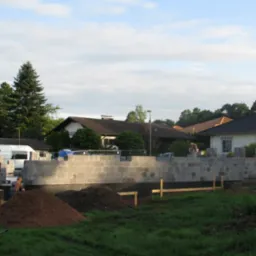 Wznoszenie ścian fundamentowych z bloczków betonowych na budowie domu jednorodzinnego, widoczne hałdy ziemi i pracownicy w trakcie murowania.