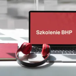 Laptop z wyświetlonym napisem 'Szkolenie BHP' na czerwonym tle, obok czerwony notatnik, słuchawki i długopis na białym stole w jasnym pomieszczeniu.
