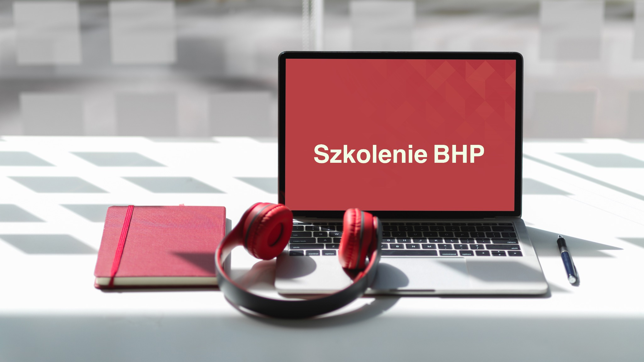 Laptop z wyświetlonym napisem 'Szkolenie BHP' na czerwonym tle, obok czerwony notatnik, słuchawki i długopis na białym stole w jasnym pomieszczeniu.
