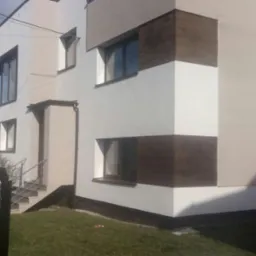 Nowoczesny dom z elewacją w odcieniach bieli, beżu i brązu, z widocznymi fragmentami o strukturze drewna, schody wejściowe z metalową barierką, widok z perspektywy trawnika.