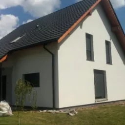 Nowoczesny dom jednorodzinny z białą elewacją, czarnym dachem i wąskimi oknami, otoczony zielonym trawnikiem, z widocznymi śladami prac wykończeniowych wokół budynku.