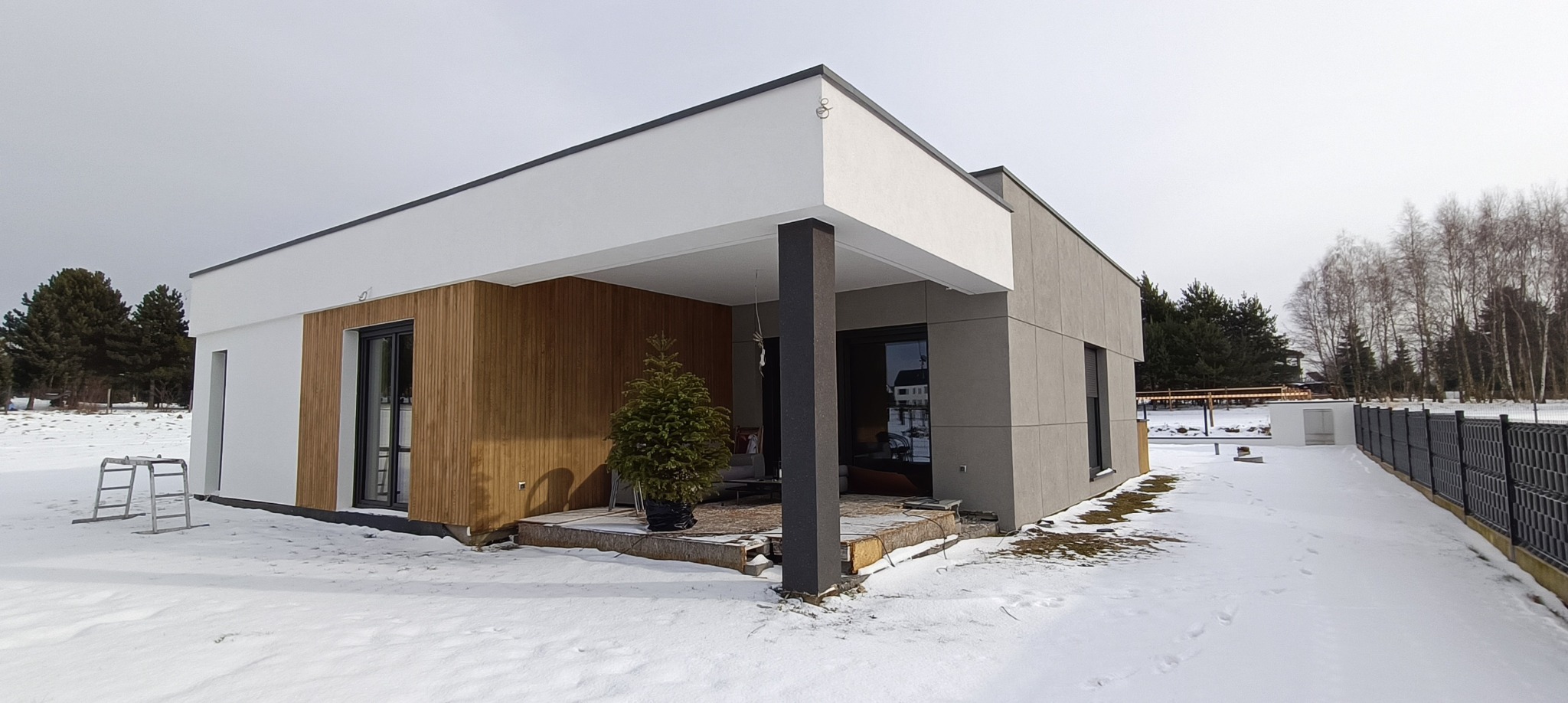 Nowoczesny dom jednorodzinny z elewacją w odcieniach bieli, szarości i drewna, częściowo pokryty śniegiem, z choinką w donicy na tarasie.