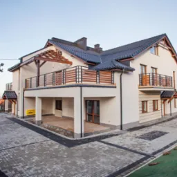 Nowoczesny, dwupiętrowy dom z balkonami i tarasem, wykończony w jasnych kolorach, z ciemną dachówką i brukowanym podjazdem z elementami dekoracyjnymi z ciemnej kostki.
