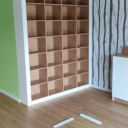 Regał z drewnianymi półkami w białej ramie, ustawiony na podłodze z paneli, z widocznymi elementami montażowymi na podłodze, na tle ściany z tapetą w motywie drzew i fragmentem zielonej ściany.