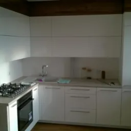 Nowoczesna kuchnia na wymiar z białymi frontami i drewnianymi akcentami. Widoczny fragment blatu z butelkami wody i płyta gazowa.