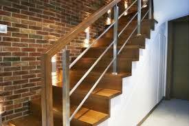 Nowoczesne schody drewniane z metalowymi balustradami, oświetlone punktowo, na tle ściany z cegły. Widok z dołu, podkreślający geometrię i materiały.