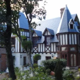 Dwukondygnacyjny dom w stylu Tudorów z elementami szachulcowymi, kryty ciemną dachówką łupkową, z wieżyczką i wysokim kominem, otoczony zielenią.