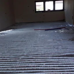 Białe rury ogrzewania podłogowego ułożone spiralnie na srebrnej folii izolacyjnej z czerwoną siatką w surowym wnętrzu budynku z oknami.