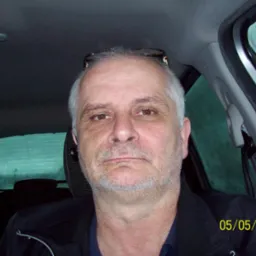 Mężczyzna z okularami na głowie, siedzący w samochodzie, patrzy w obiektyw. Widoczna data 05/05/2015. Deszcz na szybie.