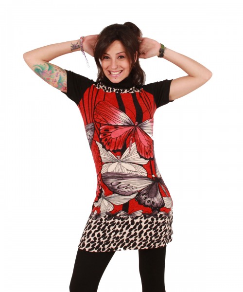 Uśmiechnięta brunetka w czerwonej tunice z motywem motyli i lampartów, czarnych legginsach, z tatuażami na ramionach, pozuje na białym tle.