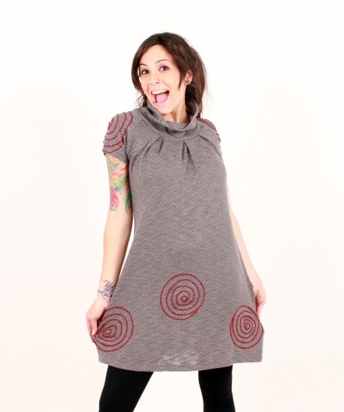 Uśmiechnięta kobieta prezentuje szarą tunikę z krótkim rękawem i czerwonymi spiralnymi wzorami, eksponując tatuaż na ramieniu.
