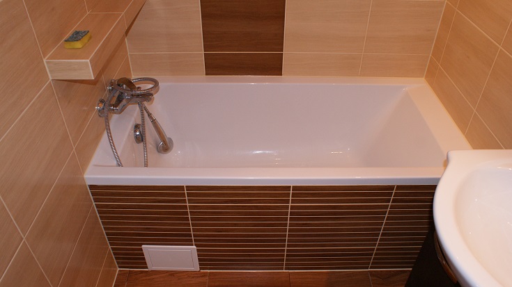 Biała wanna obudowana brązowymi płytkami imitującymi drewno, z chromowaną baterią i słuchawką prysznicową, umywalka z ceramiki w rogu, beżowe płytki na ścianach, półka z gąbką