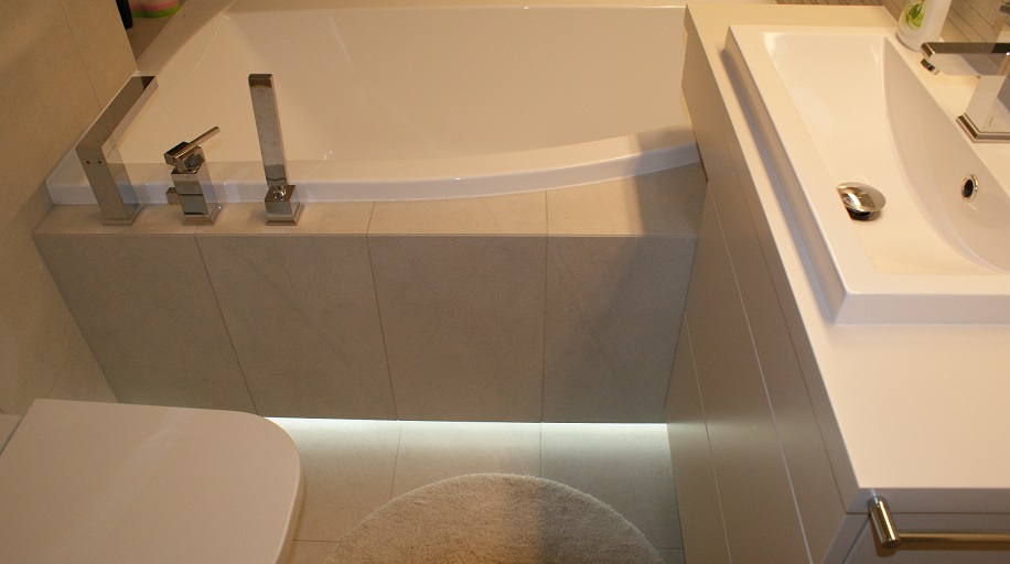 Fragment łazienki z białą wanną obudowaną płytkami, nowoczesną armaturą, umywalką wpuszczoną w blat i zaokrąglonym dywanikiem; podświetlenie LED u dołu obudowy wanny.