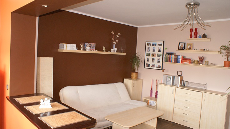 Salon po remoncie: brązowa ściana, kremowa kanapa, drewniane półki z dekoracjami, nowoczesna lampa wisząca, widok na pomarańczowy fragment ściany z boku.