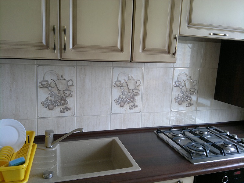 Fragment kuchni z beżowymi szafkami, brązowym blatem i ceramicznymi płytkami z motywem filiżanek, czajników i owoców, widoczny zlew z baterią i płyta gazowa.